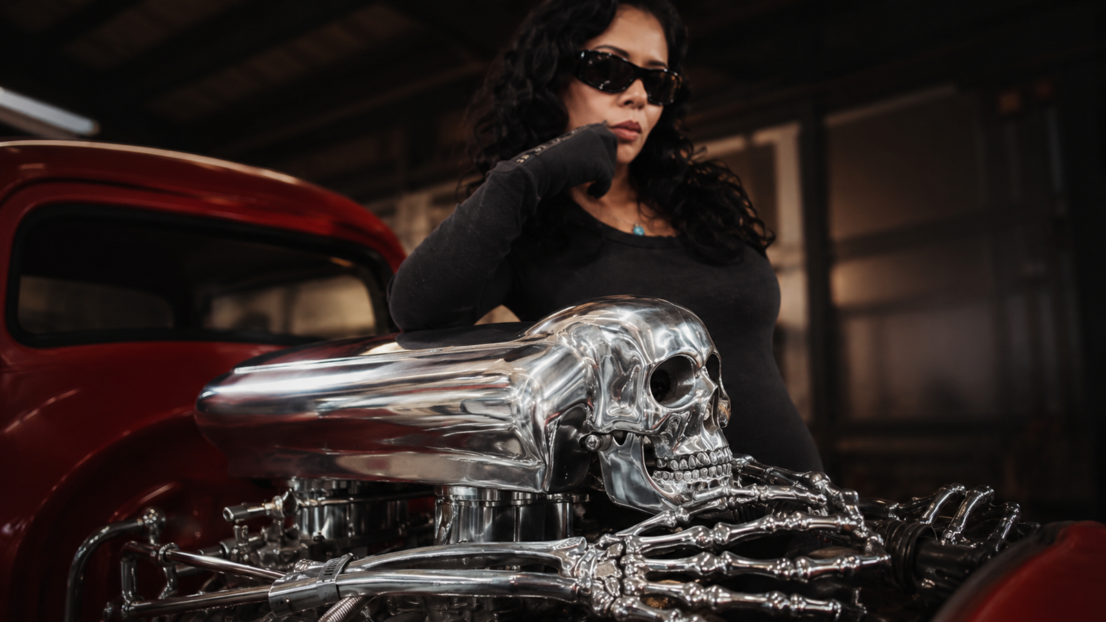 A woman with dark hair and sunglasses poses next to a red hot rod featuring a chrome skull air cleaner with skeletal hands.