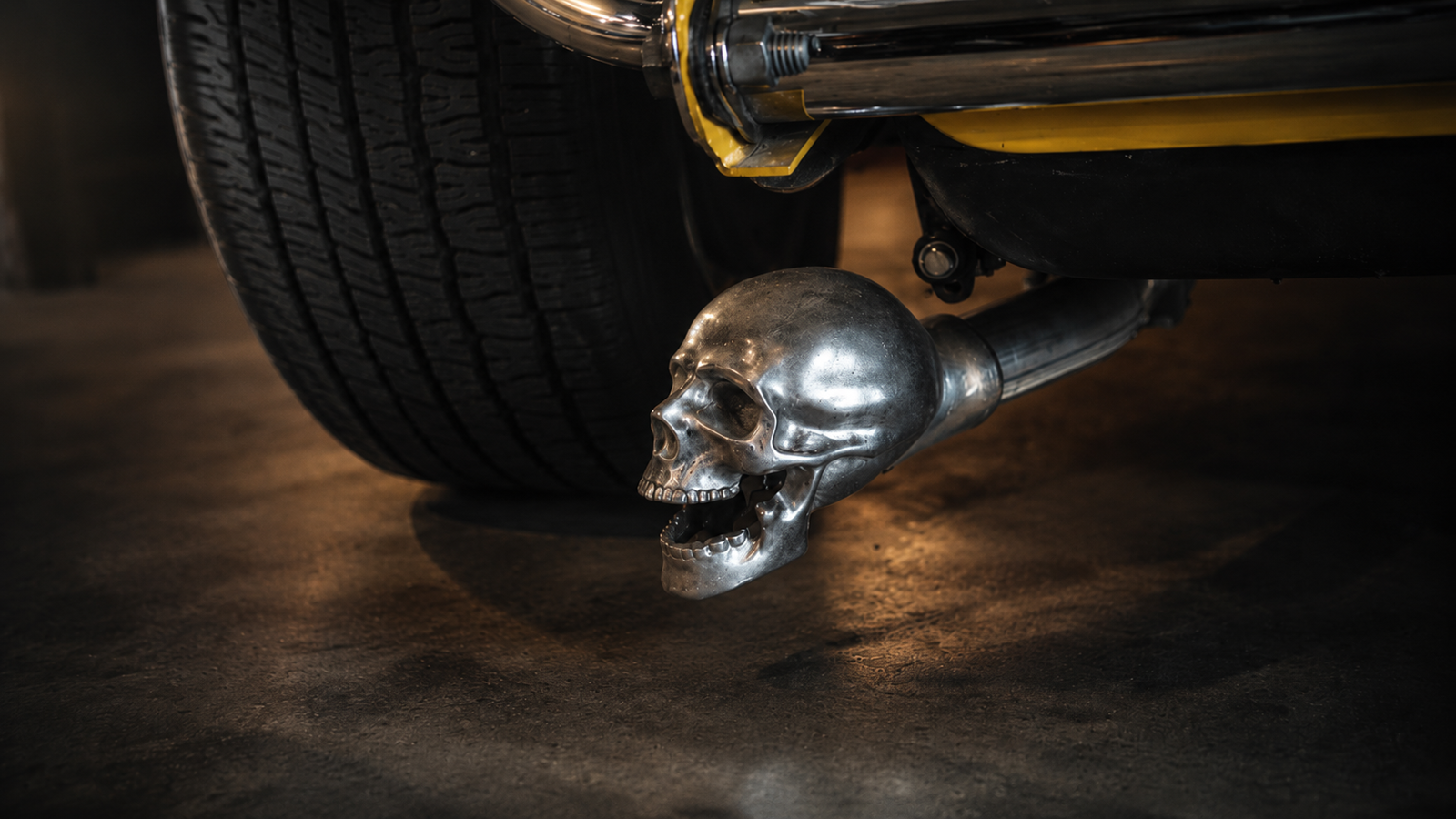 Close-up of a silver skull exhaust tip attached to a hot rod, with a large tire in the background.