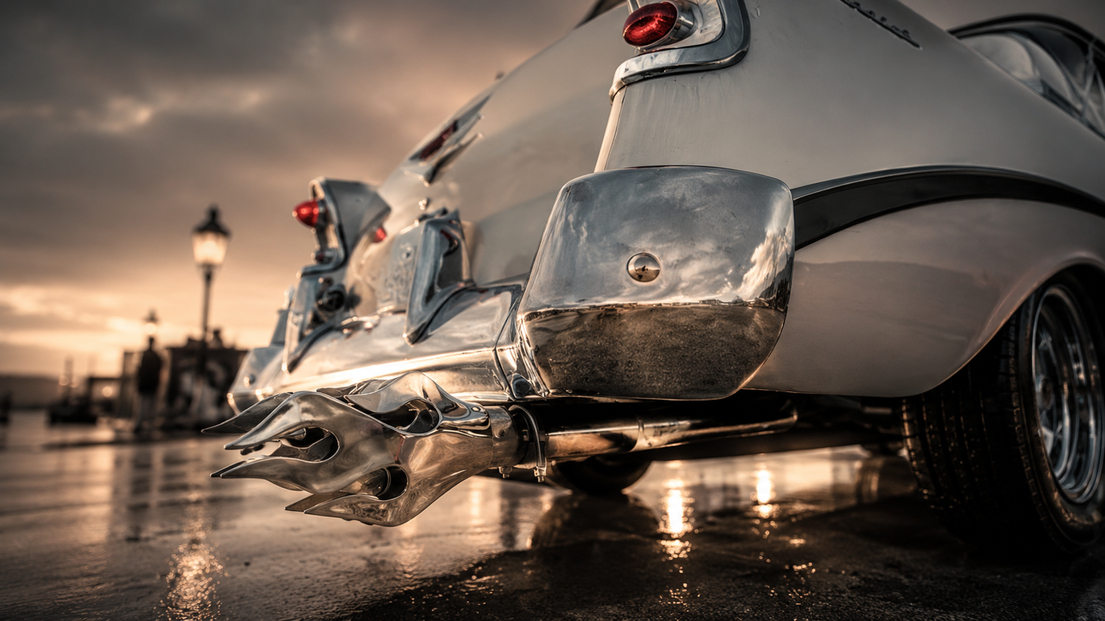 Hammer-polished chrome flame exhaust tips mounted under the rear bumper of a classic car on wet pavement at sunset.