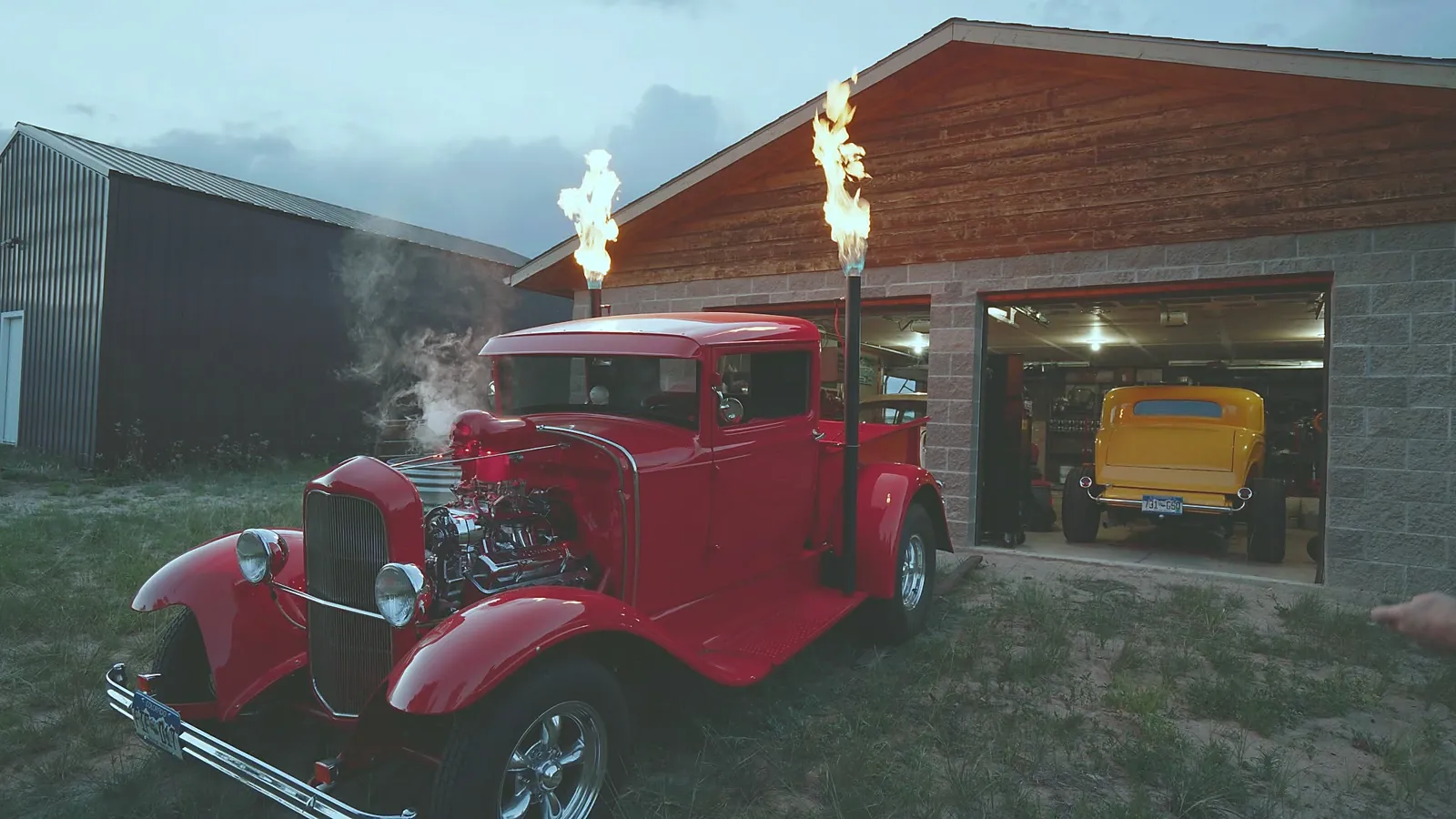Red hot rod pickup shooting flames from exhaust stacks at dusk outside a garage